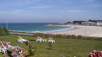 Une vue sur la plage avec des chaises longues blanches, une zone herbeuse parsemée d’hortensias et un ciel dégagé.