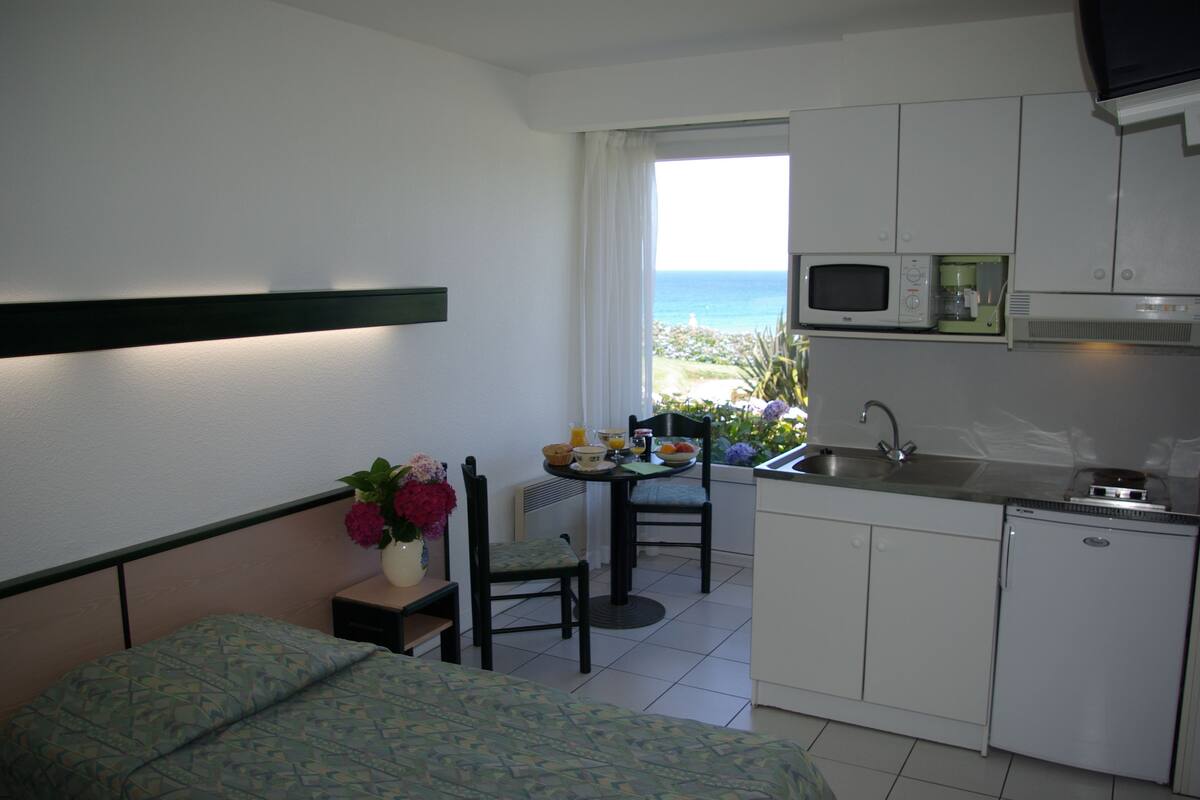 A compact hotel room with a bed, a small dining table with chairs, a kitchenette, and a view of the ocean.