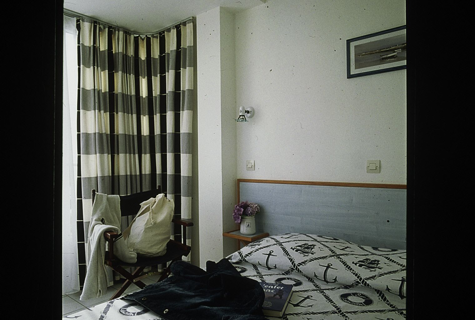 Ein Hotelzimmer mit Bett, Stuhl, kleinem Tisch und einer Vase sowie einem gestreiften Vorhang.