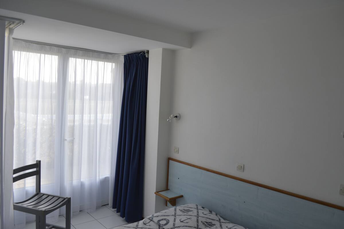 A bedroom with a bed, a chair, a small bedside table, and a window with sheer curtains and a dark blue curtain.