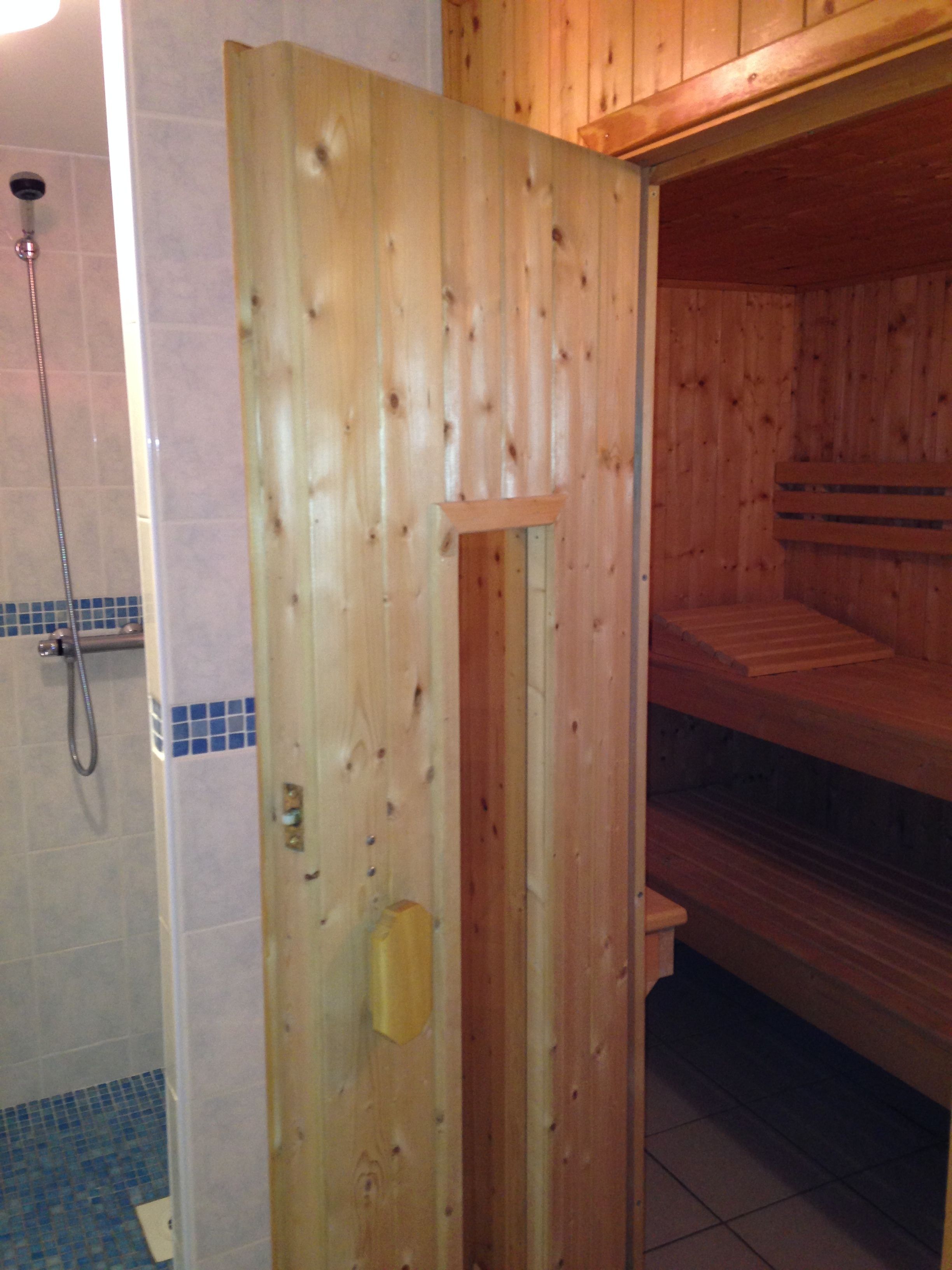 Puurakenteinen sauna, jossa on penkki, laatoitettu suihkutila ja puuovi.