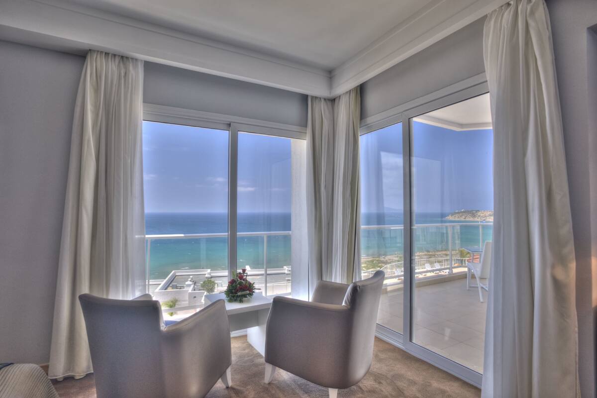 A room with a large window overlooking a coastal view, two chairs, and a small table with a flower arrangement.