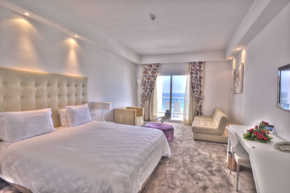A hotel room with a large bed, a sofa, a small table with a vase of flowers, and a view of the sea through the window.