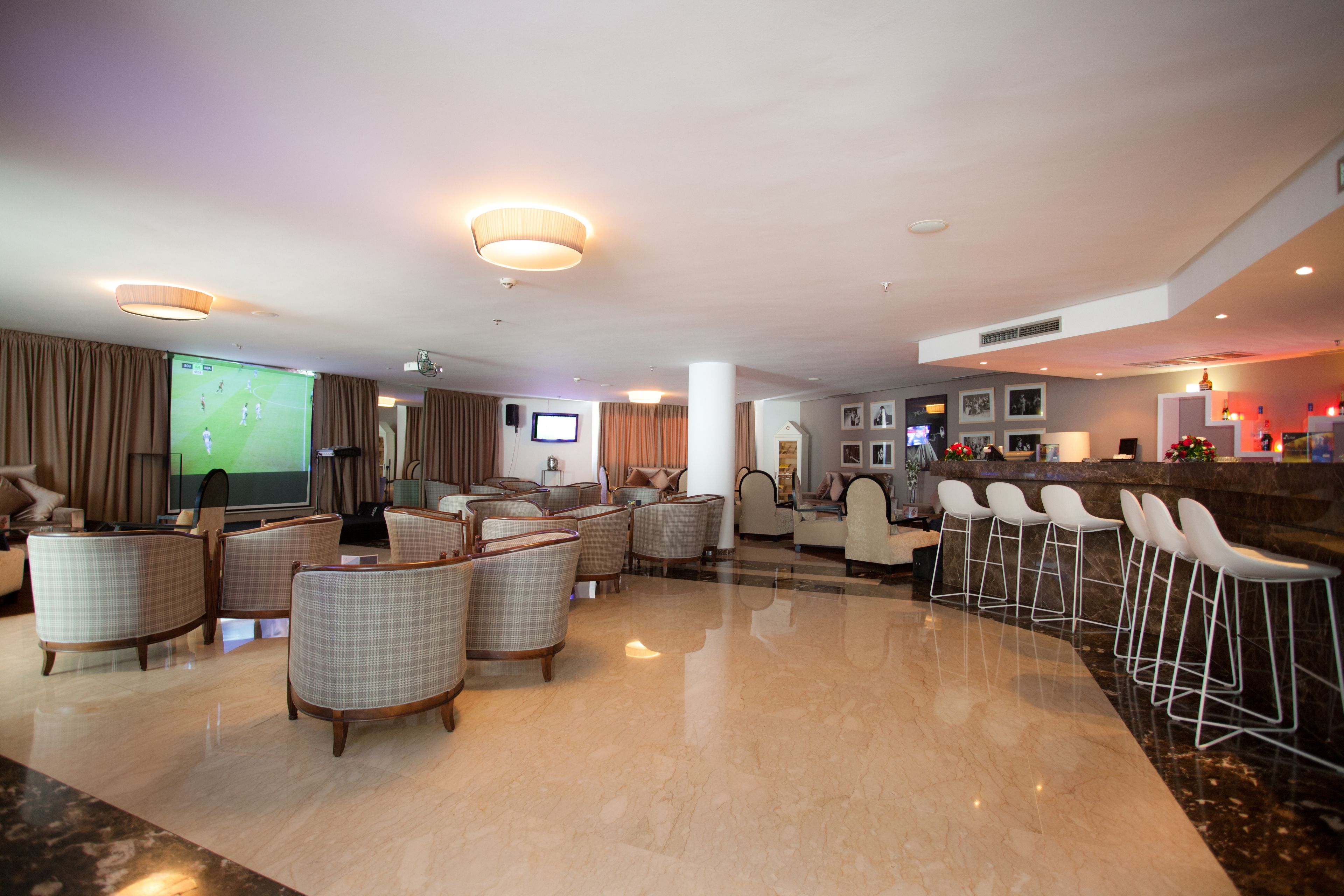 A spacious hotel lobby with a large screen displaying a soccer game, comfortable seating areas, a bar with white stools, and a reception desk.