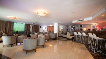 A spacious hotel lobby with a large screen displaying a soccer game, comfortable seating areas, a bar with white stools, and a reception desk.