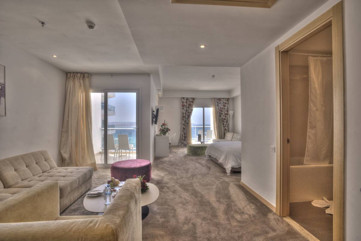 A modern hotel room with a large bed, a sofa, a coffee table, and a balcony with a view.