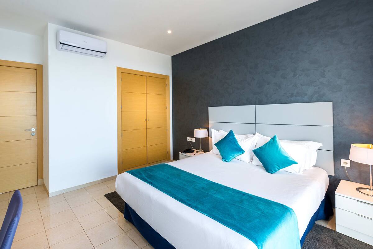 A modern hotel room with a large bed, a bedside table with a lamp, a nightstand, and a wardrobe.
