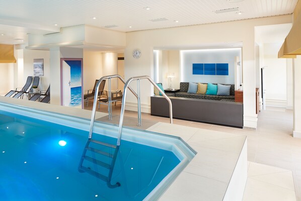 A modern indoor swimming pool with a clear view of the lounge area, a sofa, and a wall-mounted clock.