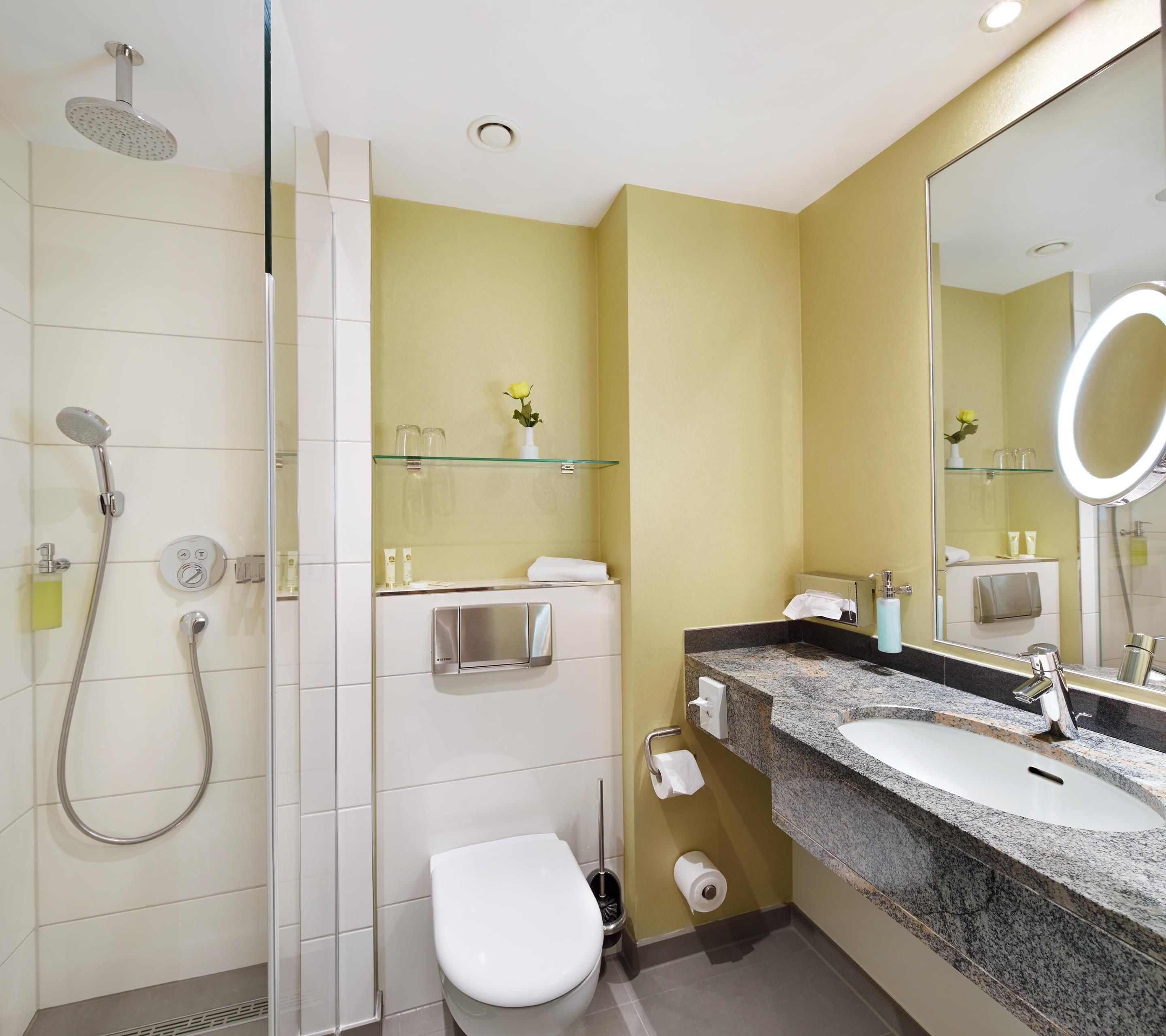 Un baño moderno con cabina de ducha de vidrio, inodoro, lavamanos con encimera de granito y espejo con luz redonda.