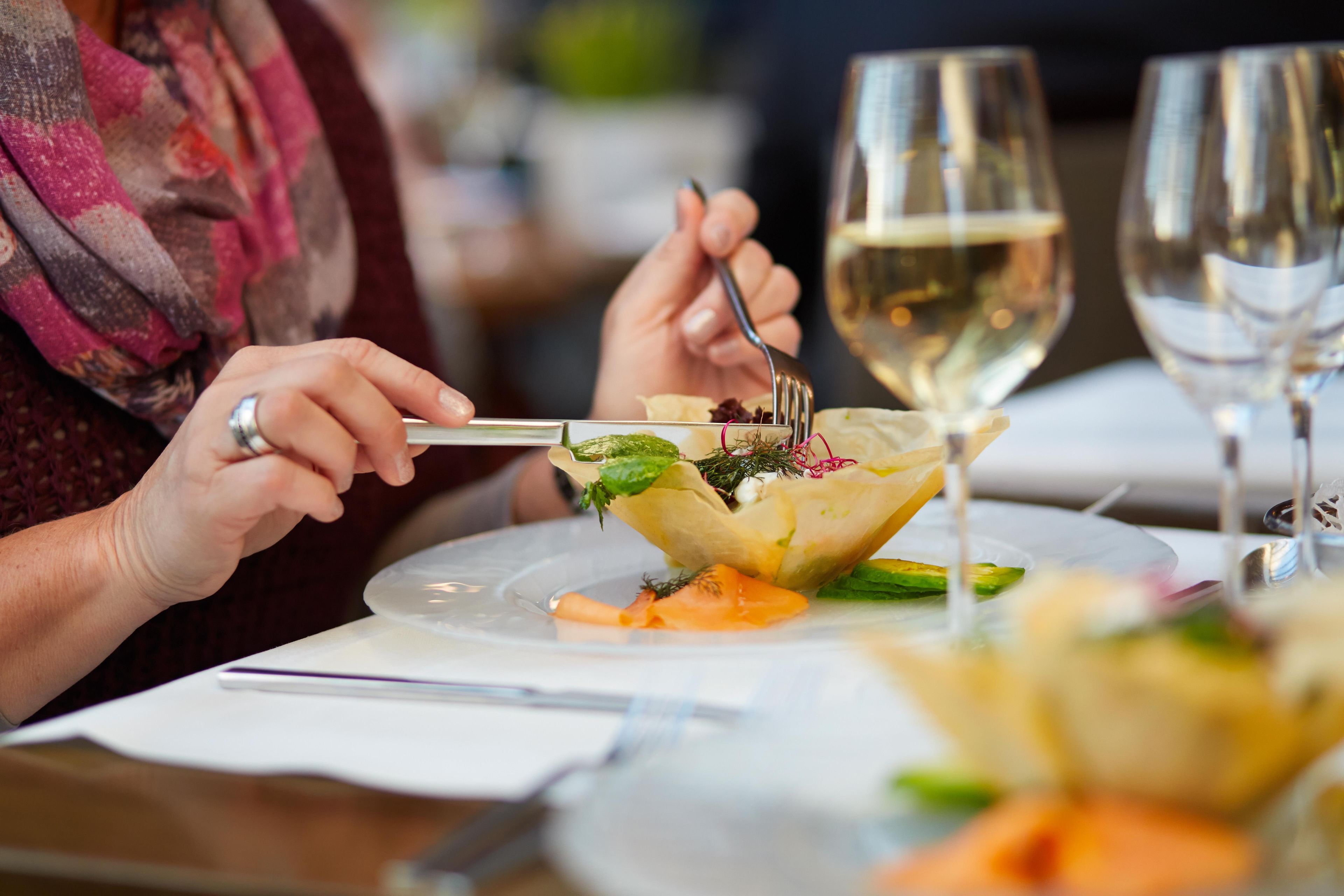 A person dining at a table with a plate of food, a fork, and a glass of white wine.