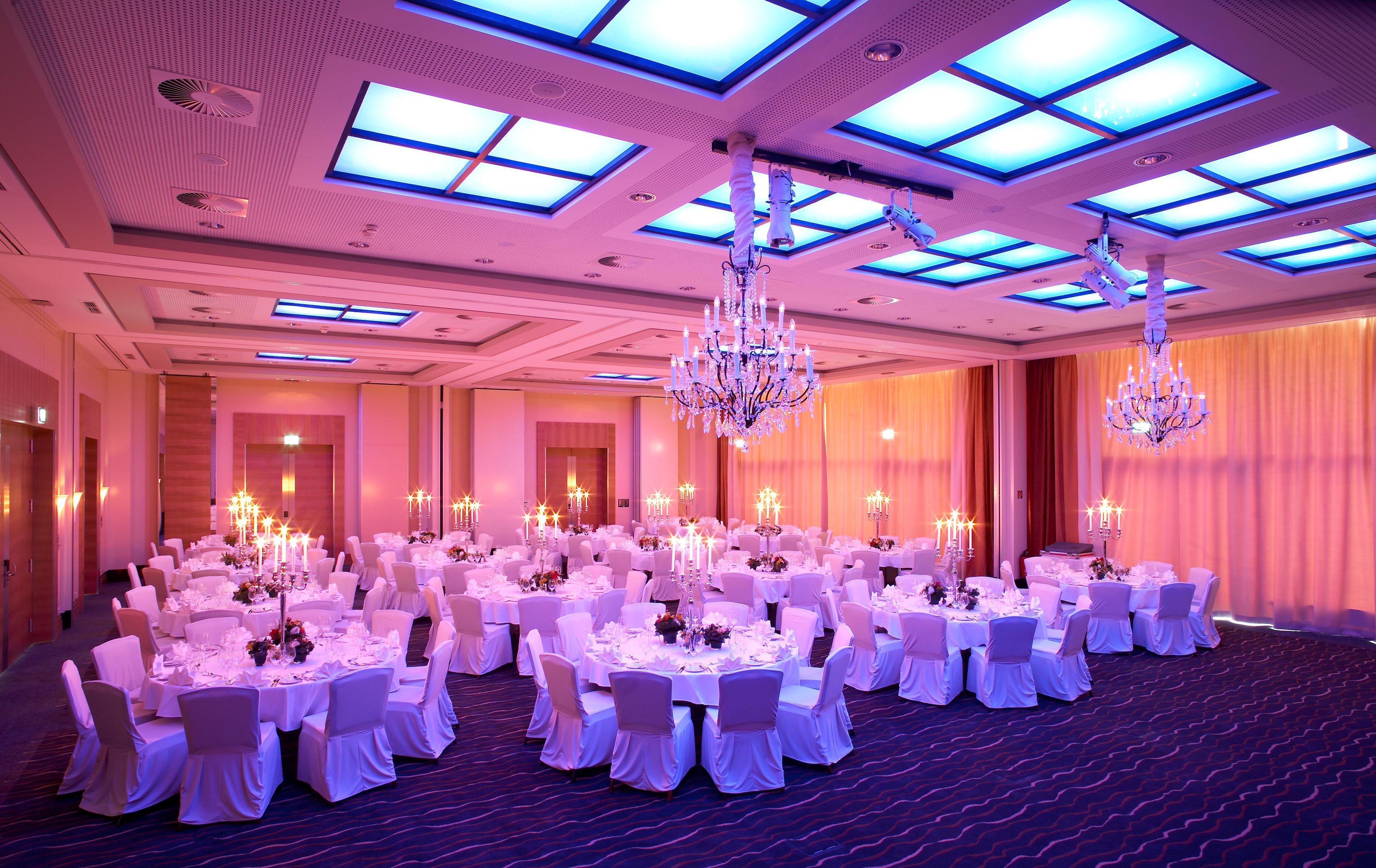 A banquet hall with round tables, white tablecloths, and chairs, illuminated by chandeliers and ceiling lights.
