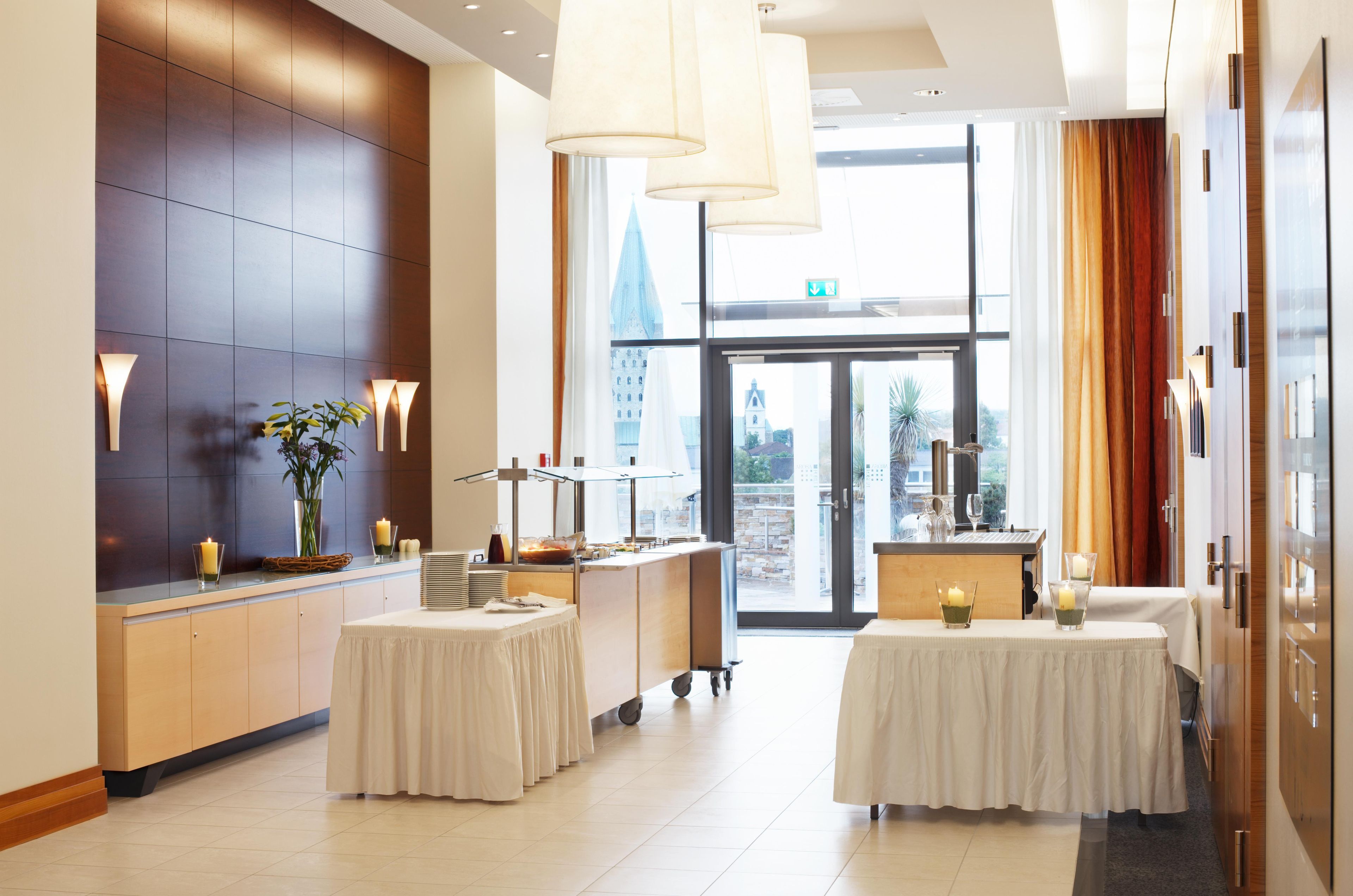 Un hall de hotel con mostrador de recepción, zona de bufé y vistas de la ciudad a través de puertas de cristal.