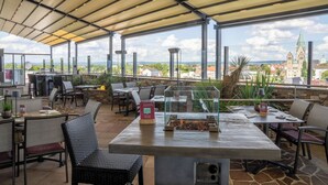 An outdoor dining area with tables and chairs, a fire feature, and a view of a town with a church. - Best Western Plus Arosa Hotel (Paderborn)