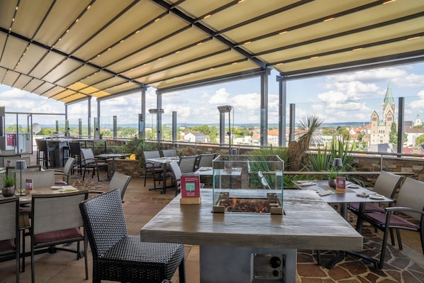 An outdoor dining area with tables and chairs, a fire feature, and a view of a town with a church. - Best Western Plus Arosa Hotel (Paderborn)
