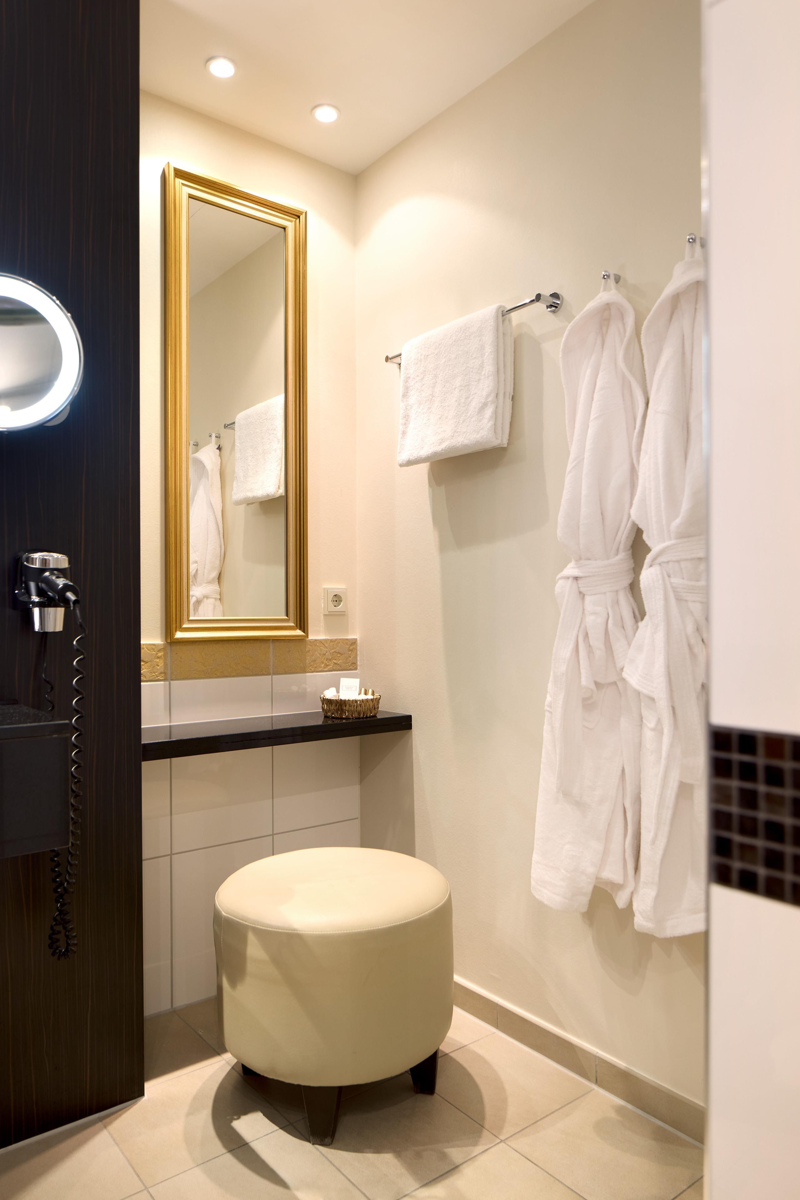 A modern bathroom with a round, cushioned stool, a large mirror with a gold frame, and two white towels on a wall-mounted rack.