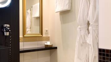 A modern bathroom with a round, cushioned stool, a large mirror with a gold frame, and two white towels on a wall-mounted rack.