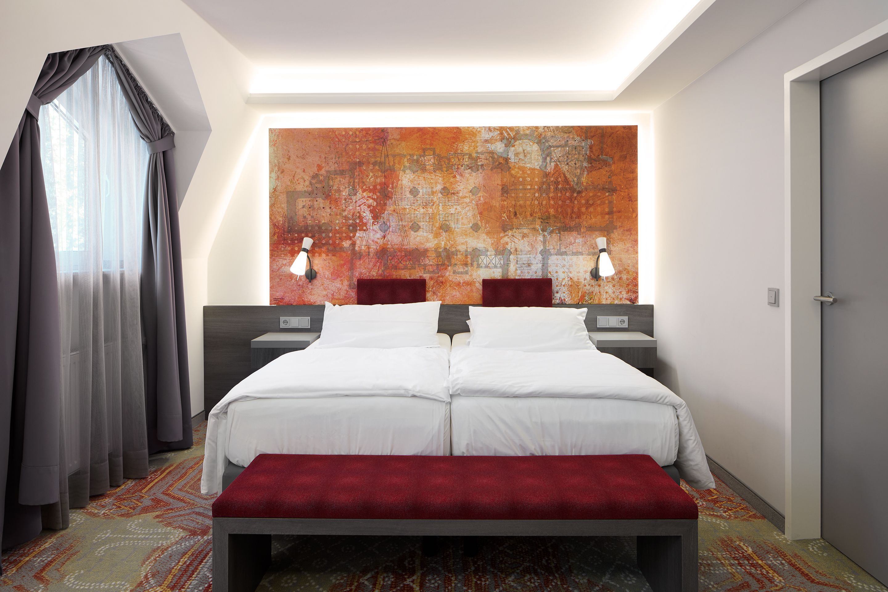 A double bed with white linens, a red bench, two bedside tables with lamps, a large abstract painting on the wall, and a patterned carpet.
