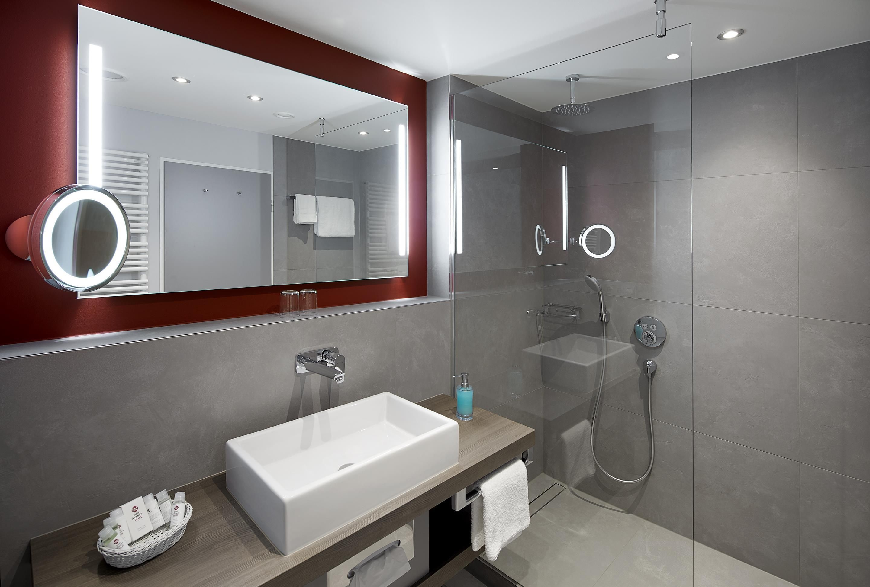 A modern bathroom with a large mirror, a white sink, a glass shower enclosure, and a red accent wall.