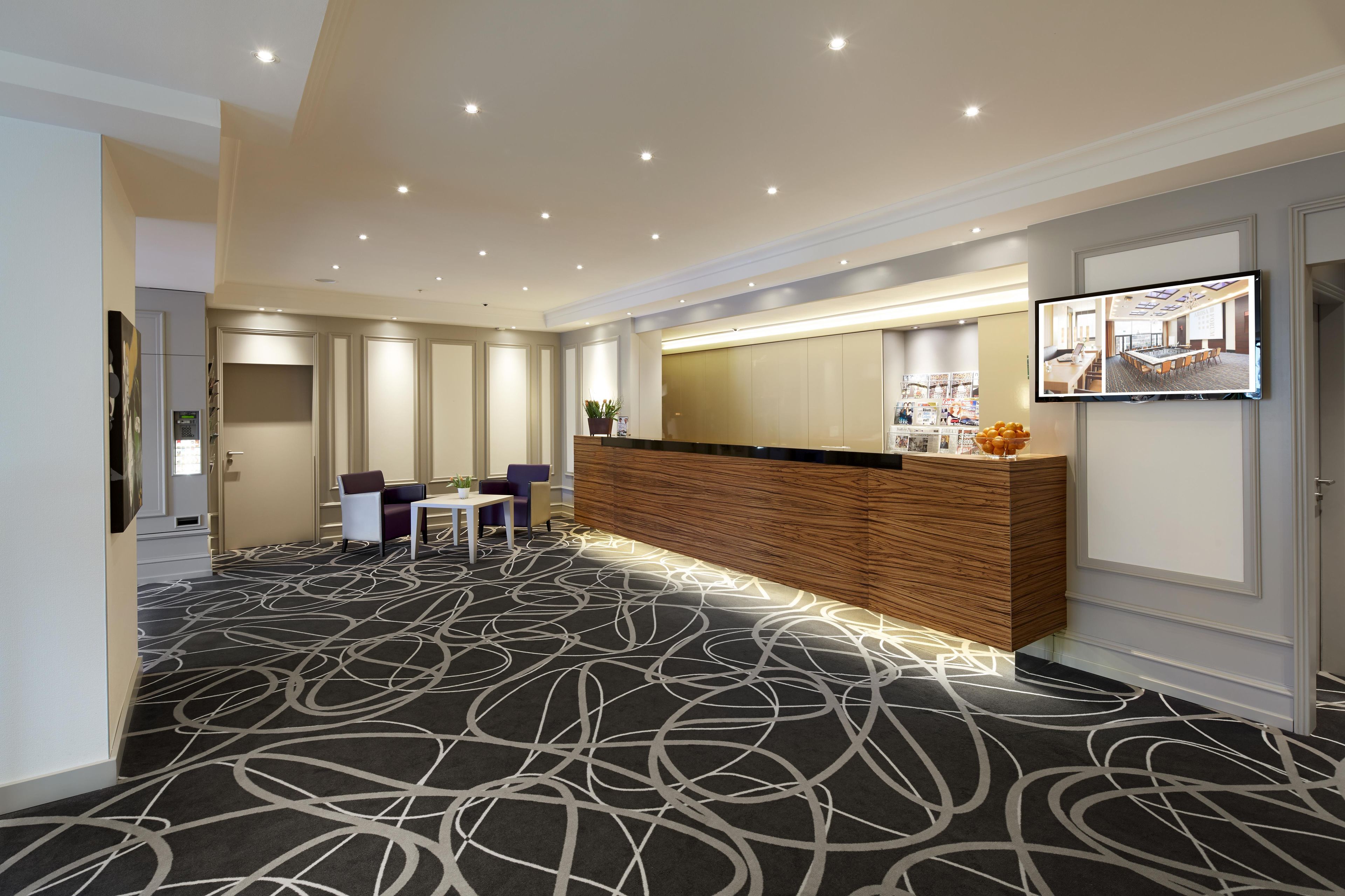 A hotel lobby with a reception desk, seating area, and a large screen displaying a room view.
