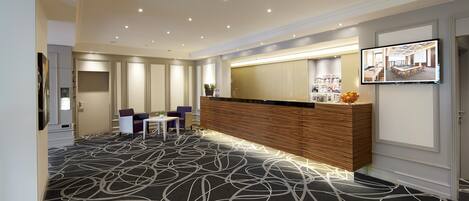 A hotel lobby with a reception desk, seating area, and a large screen displaying a room view.