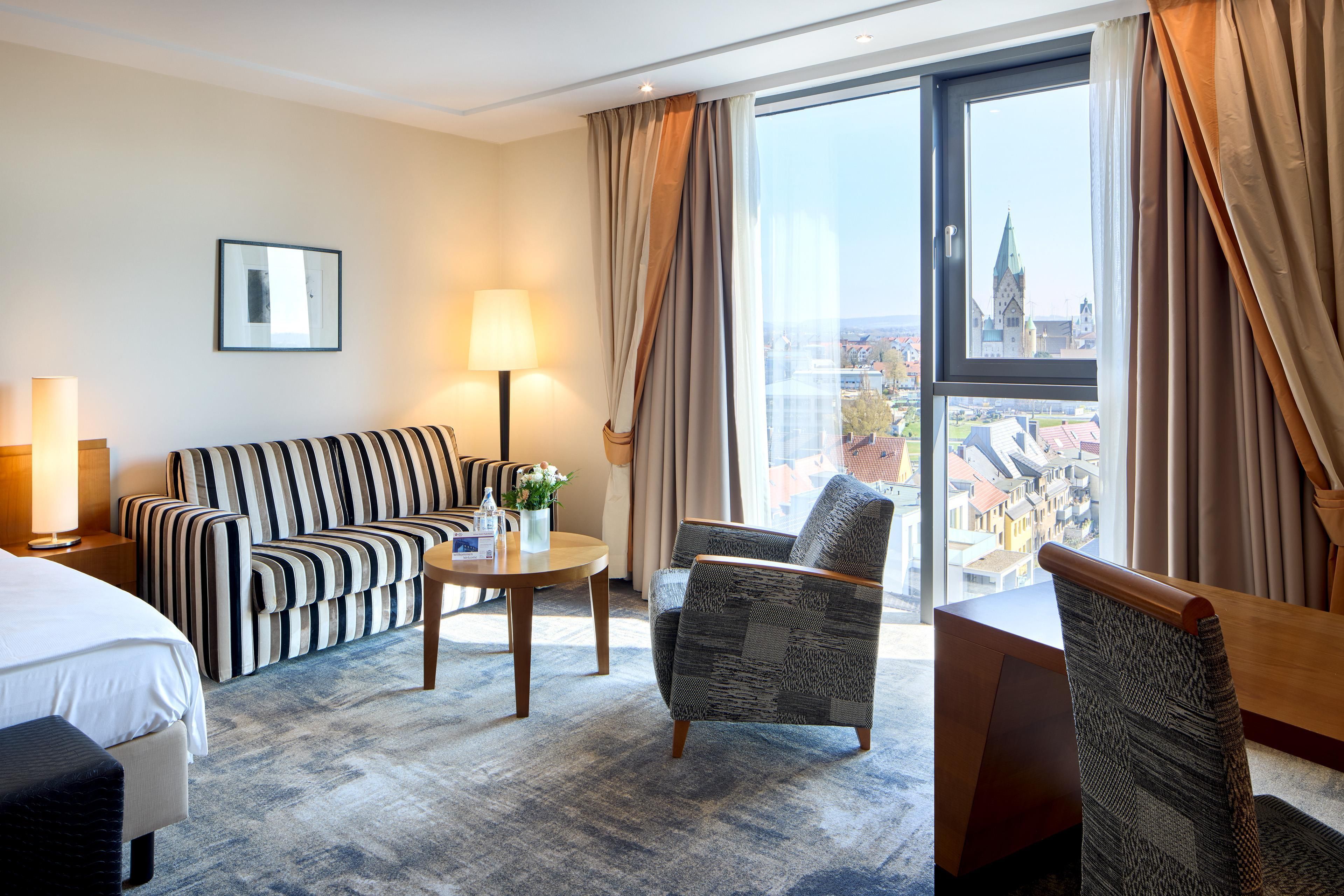 A hotel room with a bed, a striped sofa, a small table with a lamp, a chair, and a view of a cityscape through large windows.