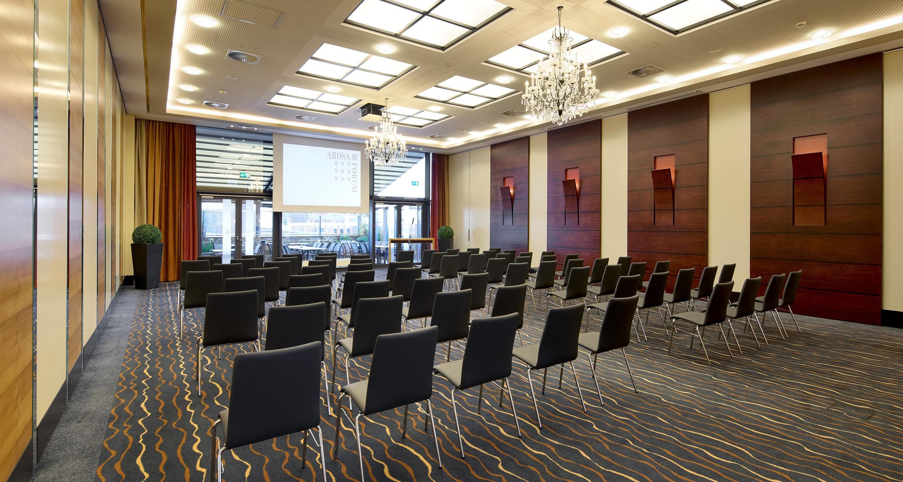 Un salón de conferencias con filas de sillas, una pantalla de proyector y un ventanal con vistas a edificios.