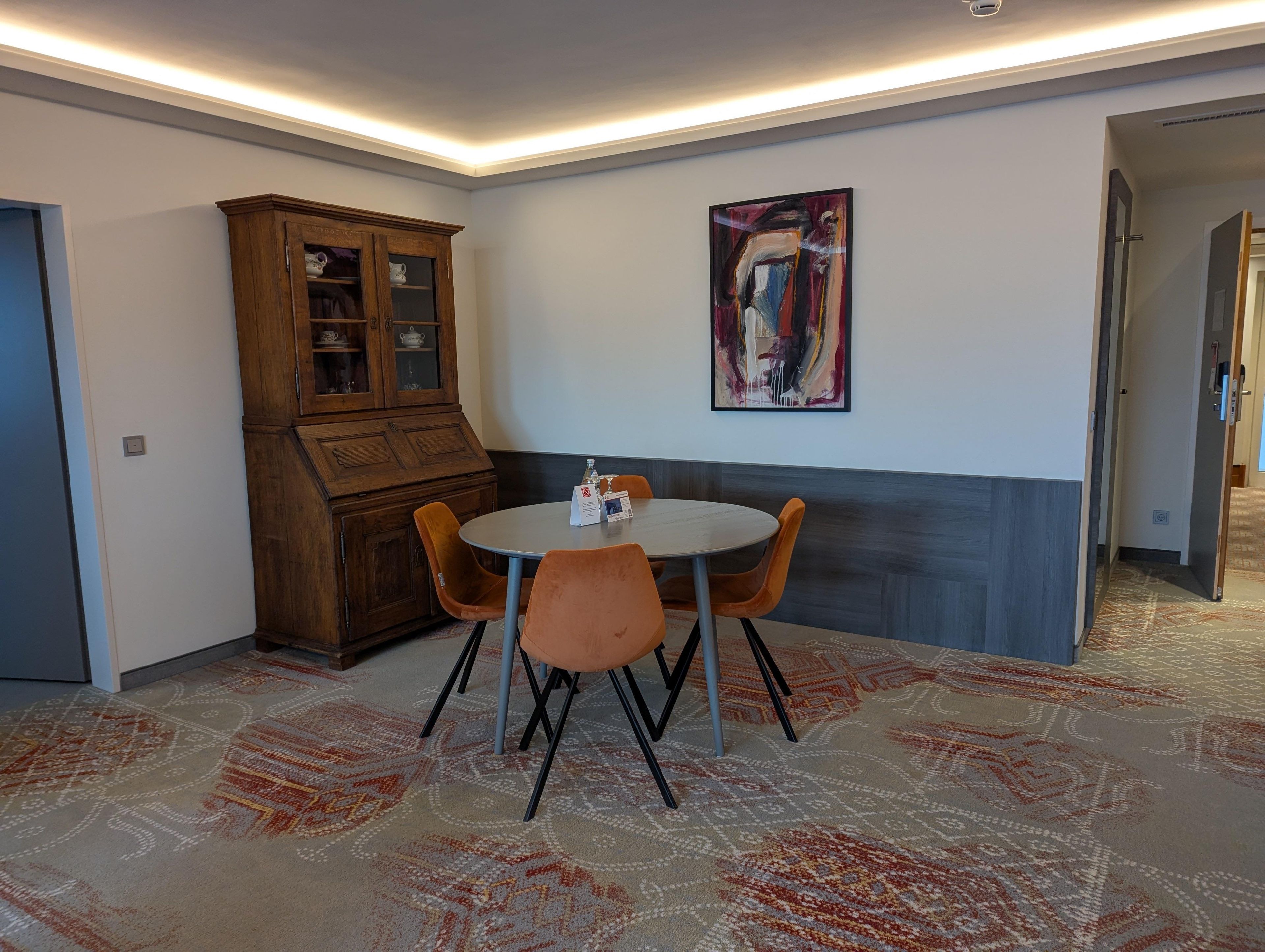 Een eethoek met een ronde tafel, oranje stoelen, een houten kast en een schilderij aan de muur.