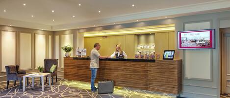 A hotel reception area with a wooden reception desk, a digital screen displaying the first floor, and a carpeted floor with a geometric pattern.