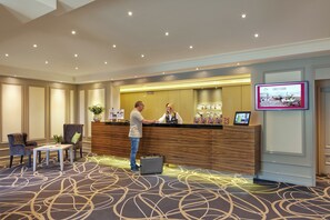 A hotel reception area with a wooden reception desk, a digital screen displaying the first floor, and a carpeted floor with a geometric pattern. - Best Western Plus Arosa Hotel (Paderborn)
