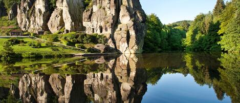 Formación rocosa natural con un reflejo nítido en el agua, rodeada de vegetación y un pequeño edificio.