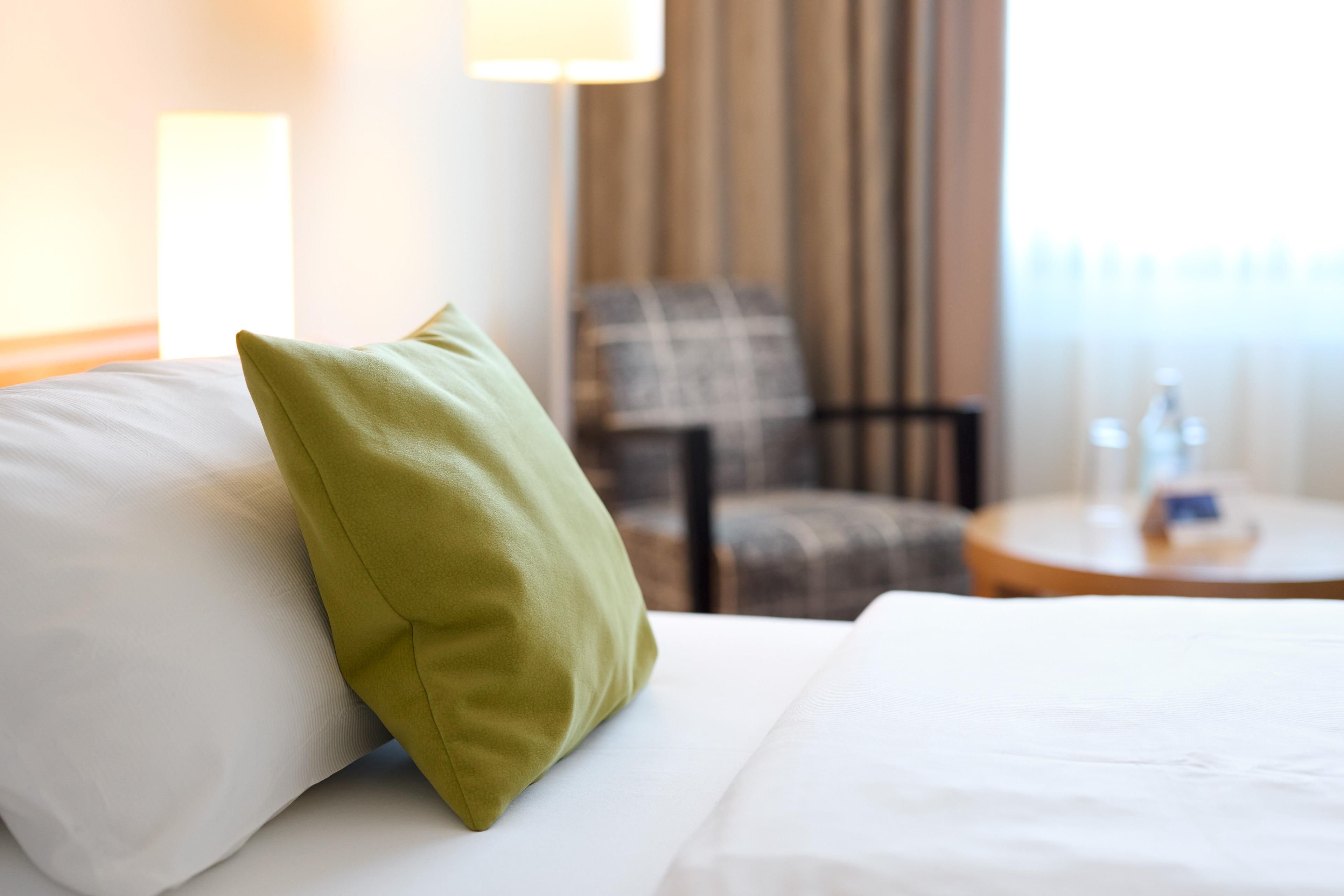A hotel room with a bed, a pillow, a chair, and a round table with a bottle on it.