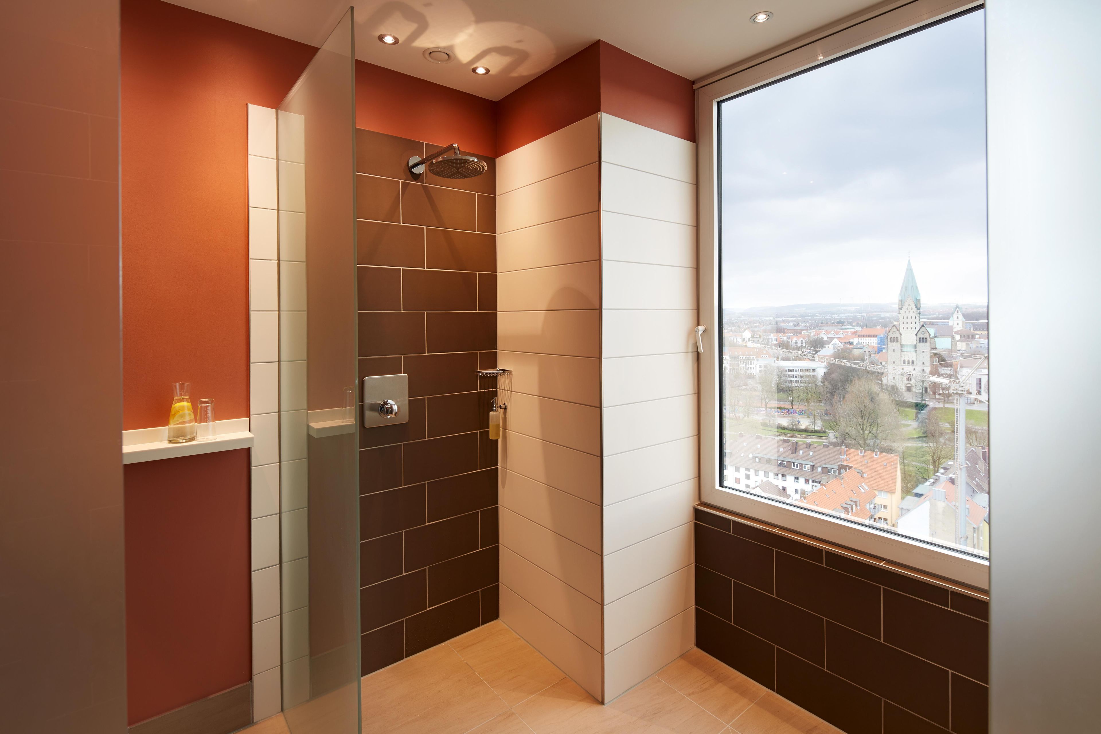 A modern shower with a glass door, a view of a cityscape through a large window, and a shelf with toiletries.