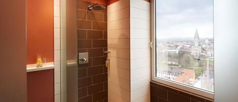 A modern shower with a glass door, a view of a cityscape through a large window, and a shelf with toiletries.