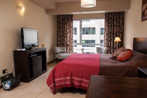Une chambre d’hôtel avec un lit, un téléviseur posé sur une commode, une petite table avec des chaises et une vue sur un immeuble par la fenêtre.