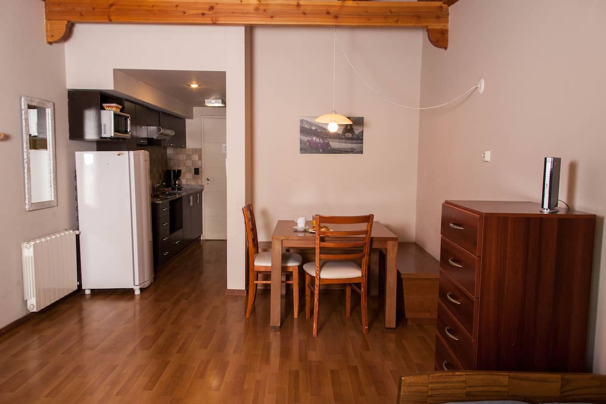 Cocina compacta con refrigerador, microondas y armarios de madera. Se ve una mesa de comedor con sillas y un mueble de madera con cajones.