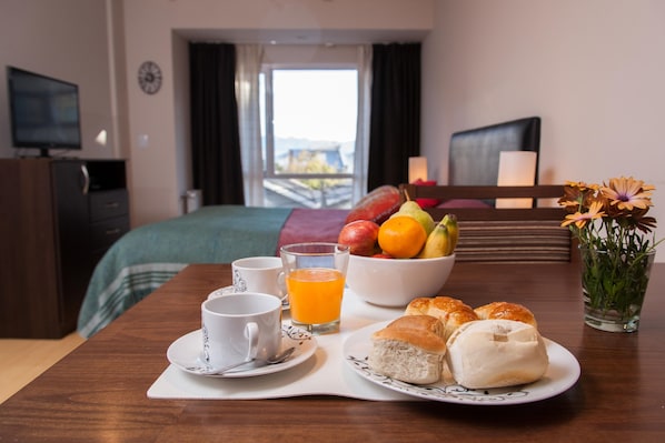 A hotel room with a bed, a television, a breakfast table with a cup, a glass of orange juice, a bowl of fruit, and a vase with flowers.