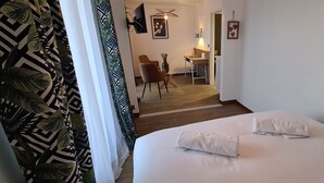Une chambre d’hôtel avec un lit, deux serviettes, une chaise, une petite table, un tableau au mur et une plante.