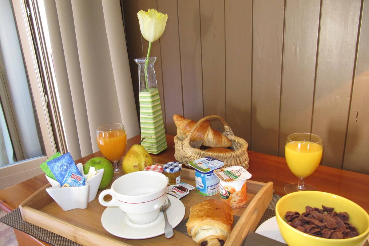 Un plateau de petit-déjeuner avec des croissants, une poire, un verre de jus d’orange, un bol de céréales, un petit vase avec une fleur et un pot de yaourt.