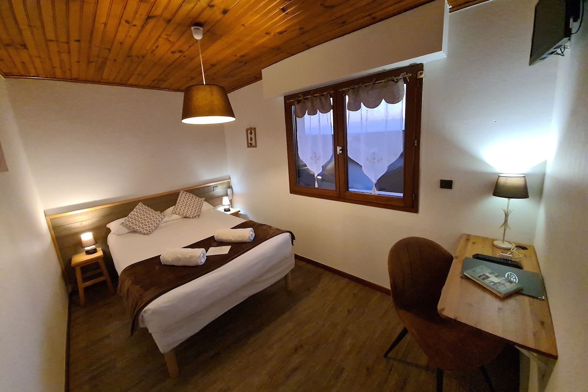 Une chambre avec un plafond en bois, un lit recouvert de draps blancs, une table de chevet avec une lampe, une fenêtre avec des rideaux et un bureau avec une chaise.