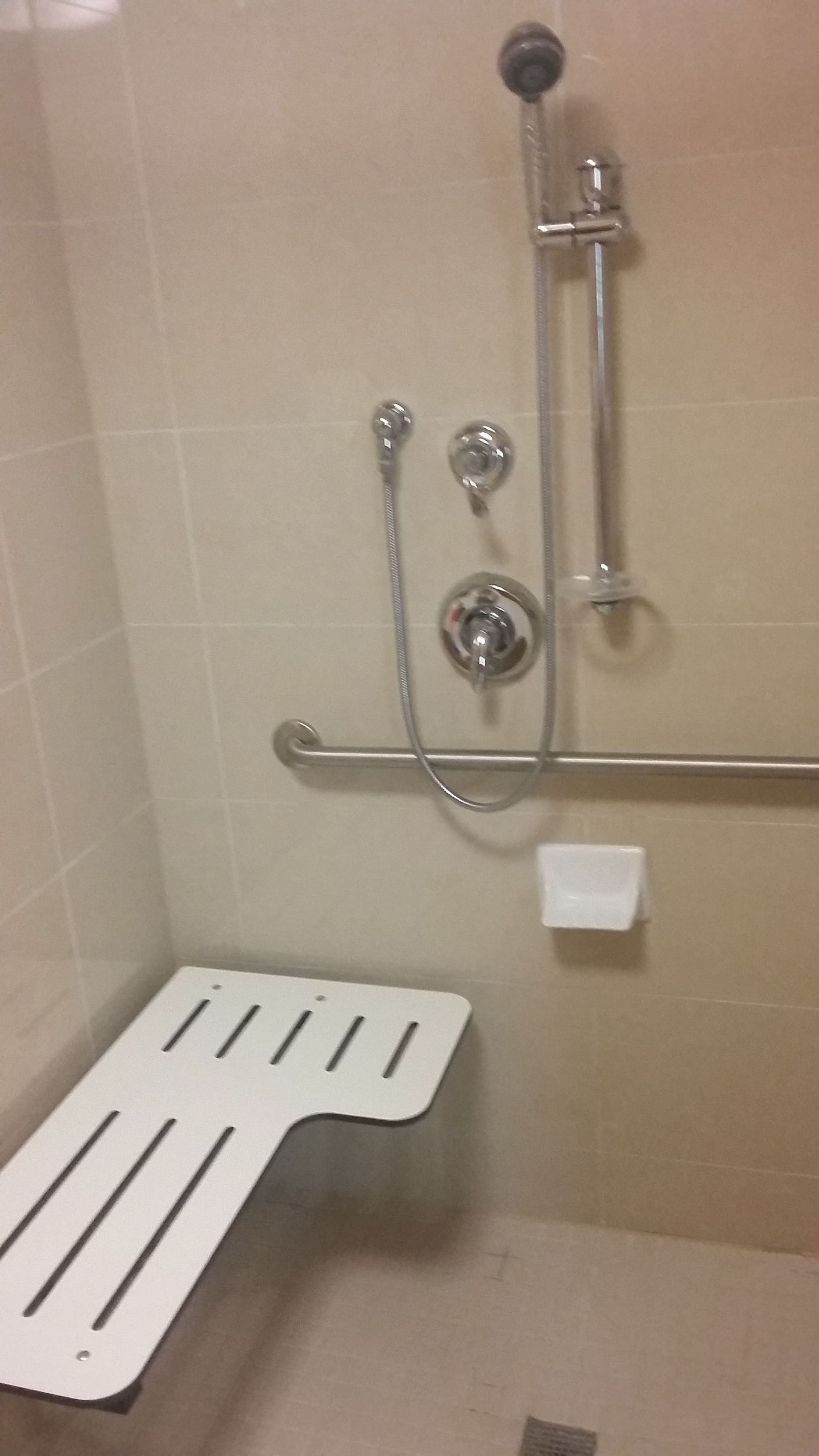 Área de chuveiro com chuveiro manual, torneira fixada na parede e barra de apoio.