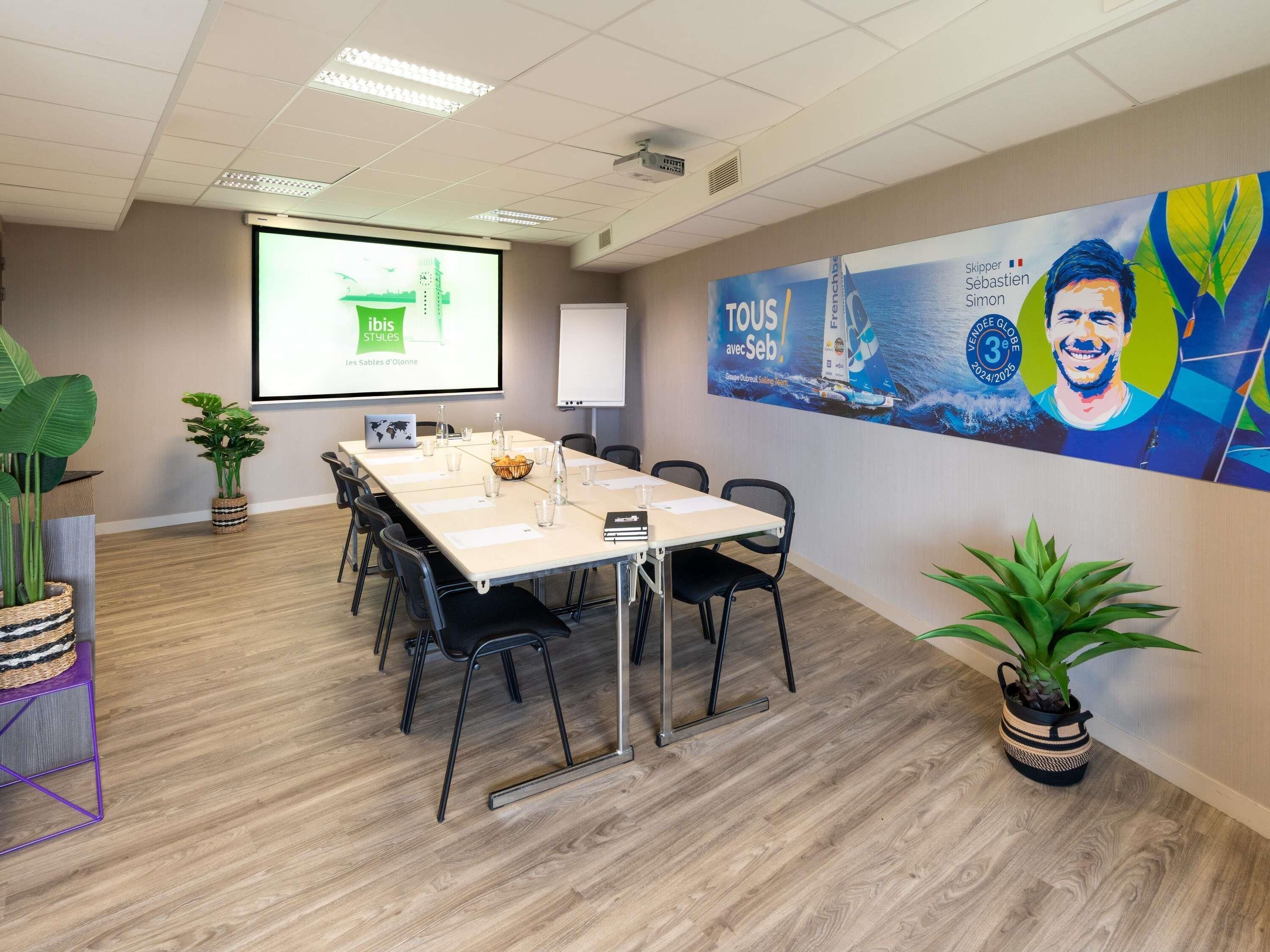 A conference room with a long table, chairs, a projector screen, and a large poster on the wall.