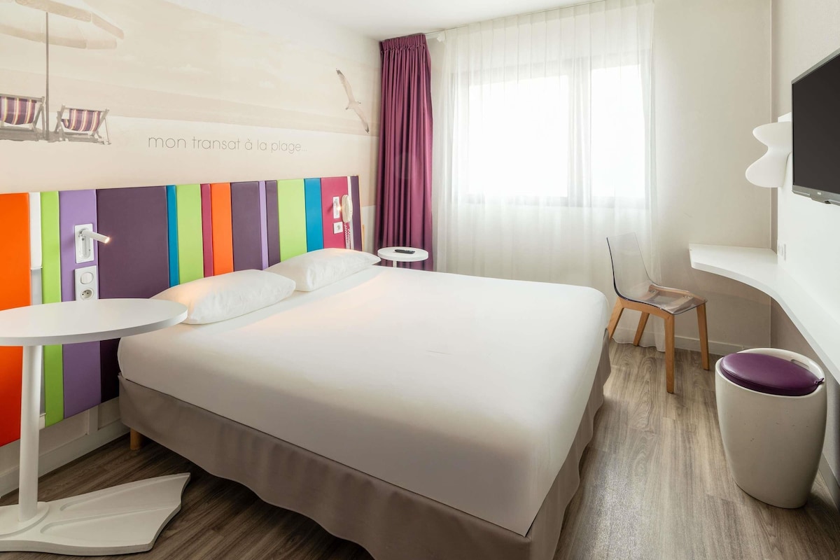 A hotel room with a bed, a bedside table, a chair, a TV, and a window with curtains.