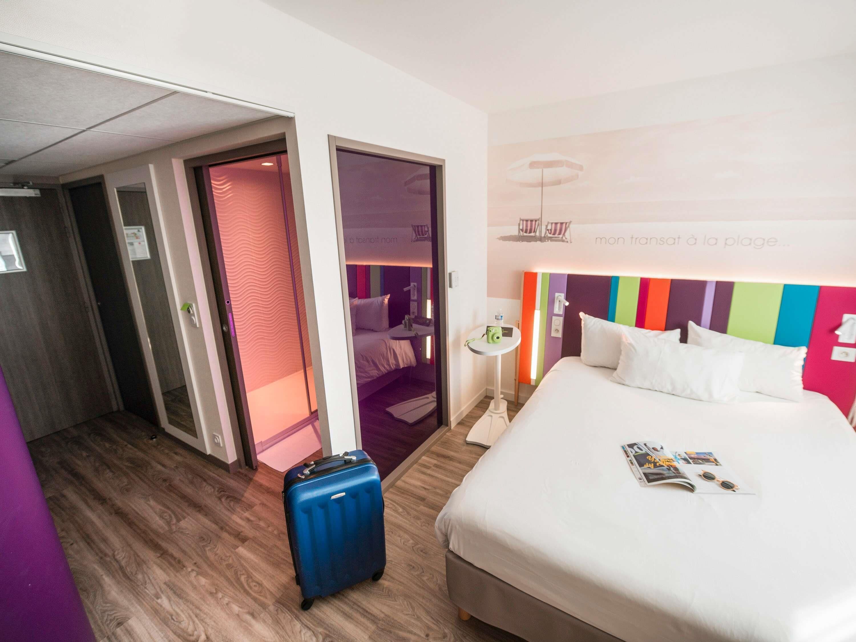 Una habitación de hotel con una cama, una maleta, una mesita con una lámpara y una pared con un estampado a rayas de colores.