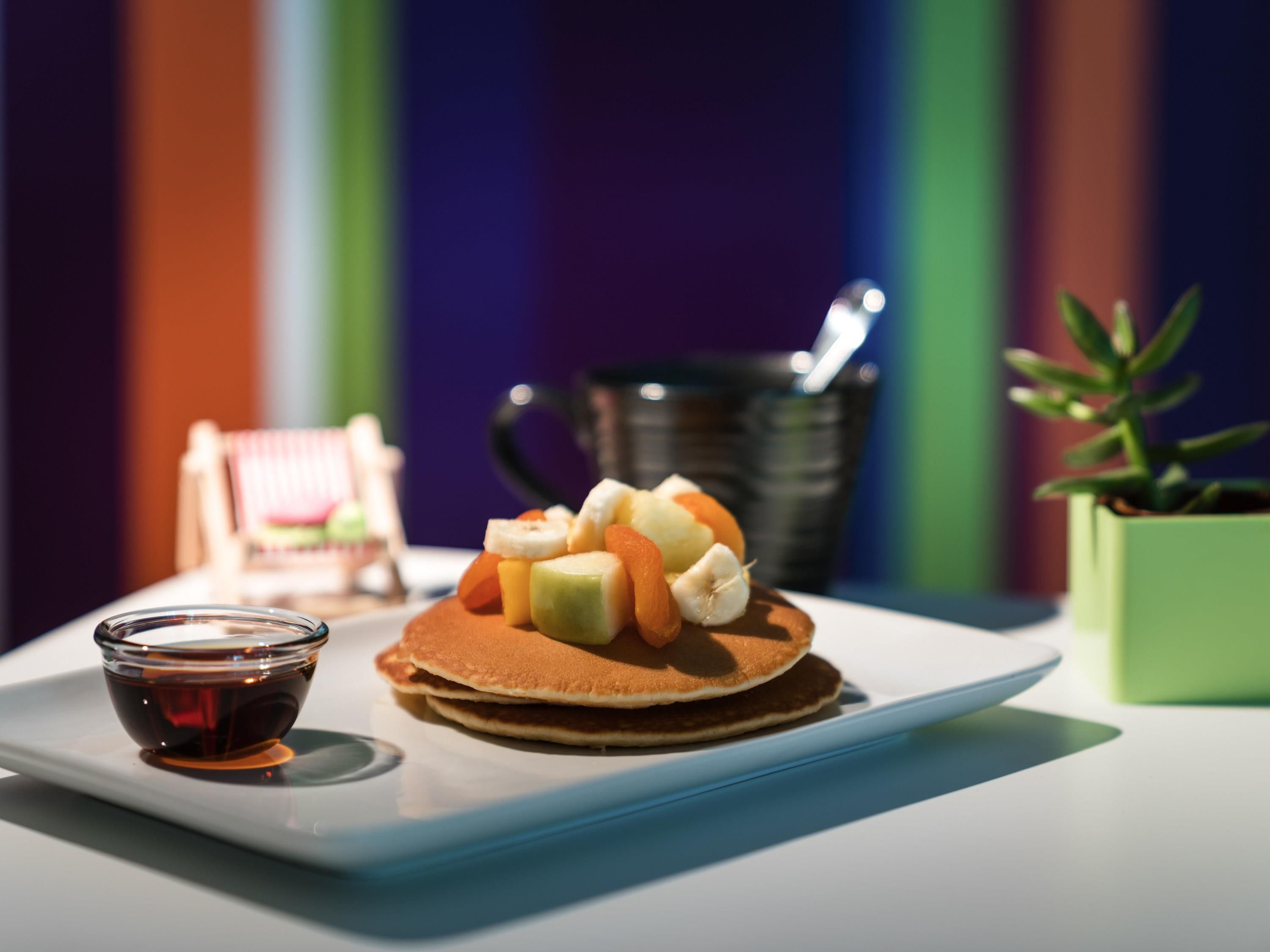 Un plato de desayuno con panqueques y fruta, una taza pequeña de almíbar y una cafetera al fondo.