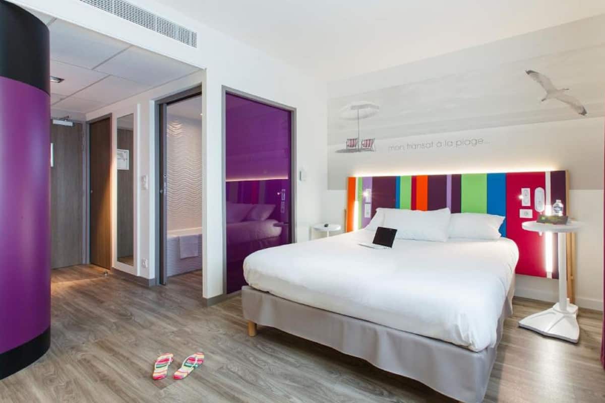 Una habitación de hotel moderna con una cama grande, una mesita de noche y una pequeña mesa auxiliar. Se ve una puerta que da a otra habitación y una ventana con cortinas.