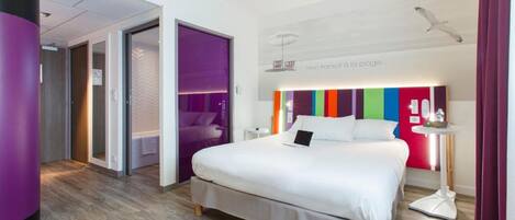 Una habitación de hotel moderna con una cama grande, una mesita de noche y una pequeña mesa auxiliar. Se ve una puerta que da a otra habitación y una ventana con cortinas.