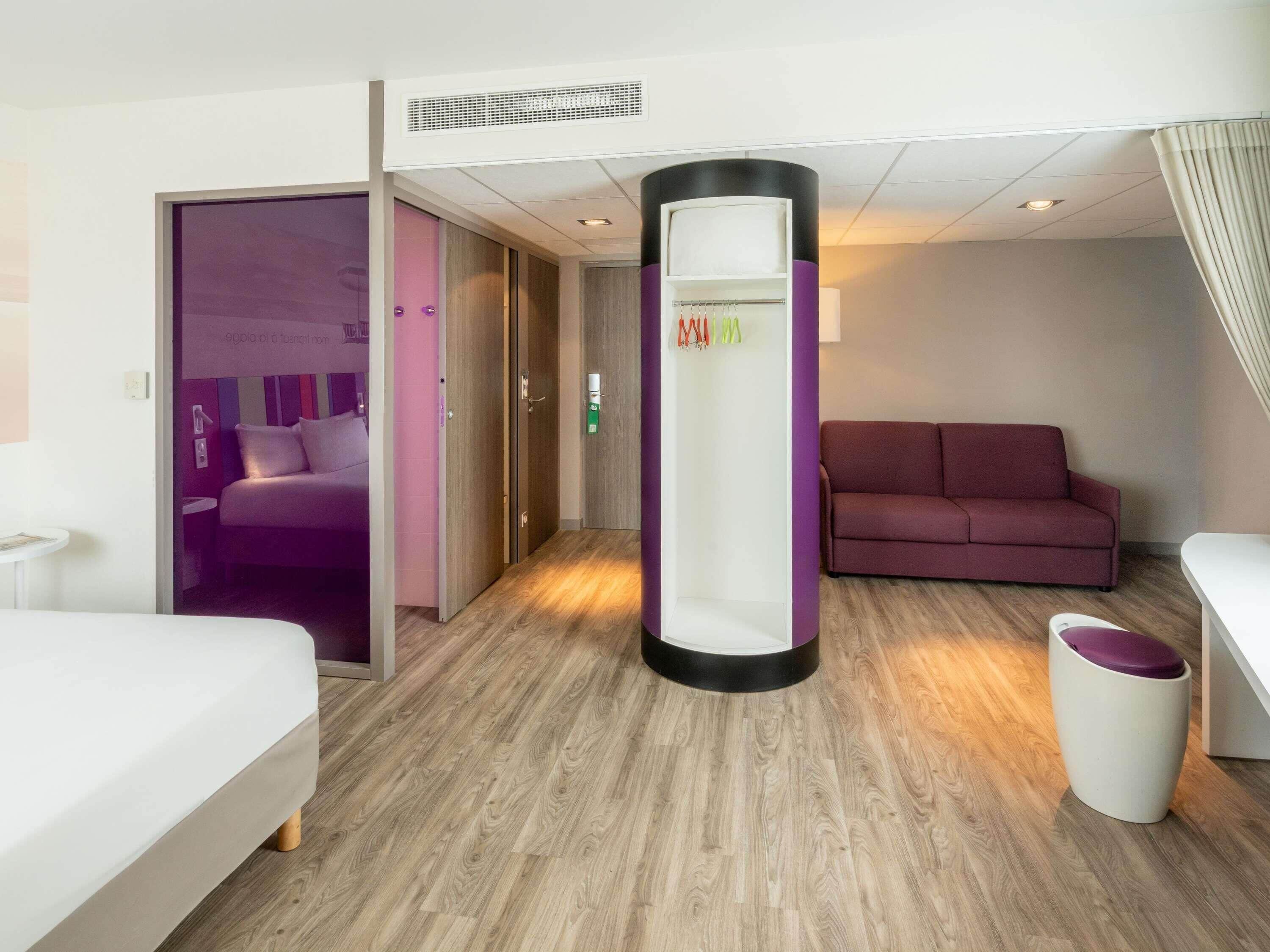 A hotel room with a bed, a sofa, a small table, a trash bin, and a bathroom with a shower.