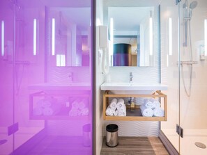 Un baño moderno con cabina de ducha de vidrio, un lavabo blanco y un espejo sobre él. Toallas están ordenadas en un estante debajo del lavabo.