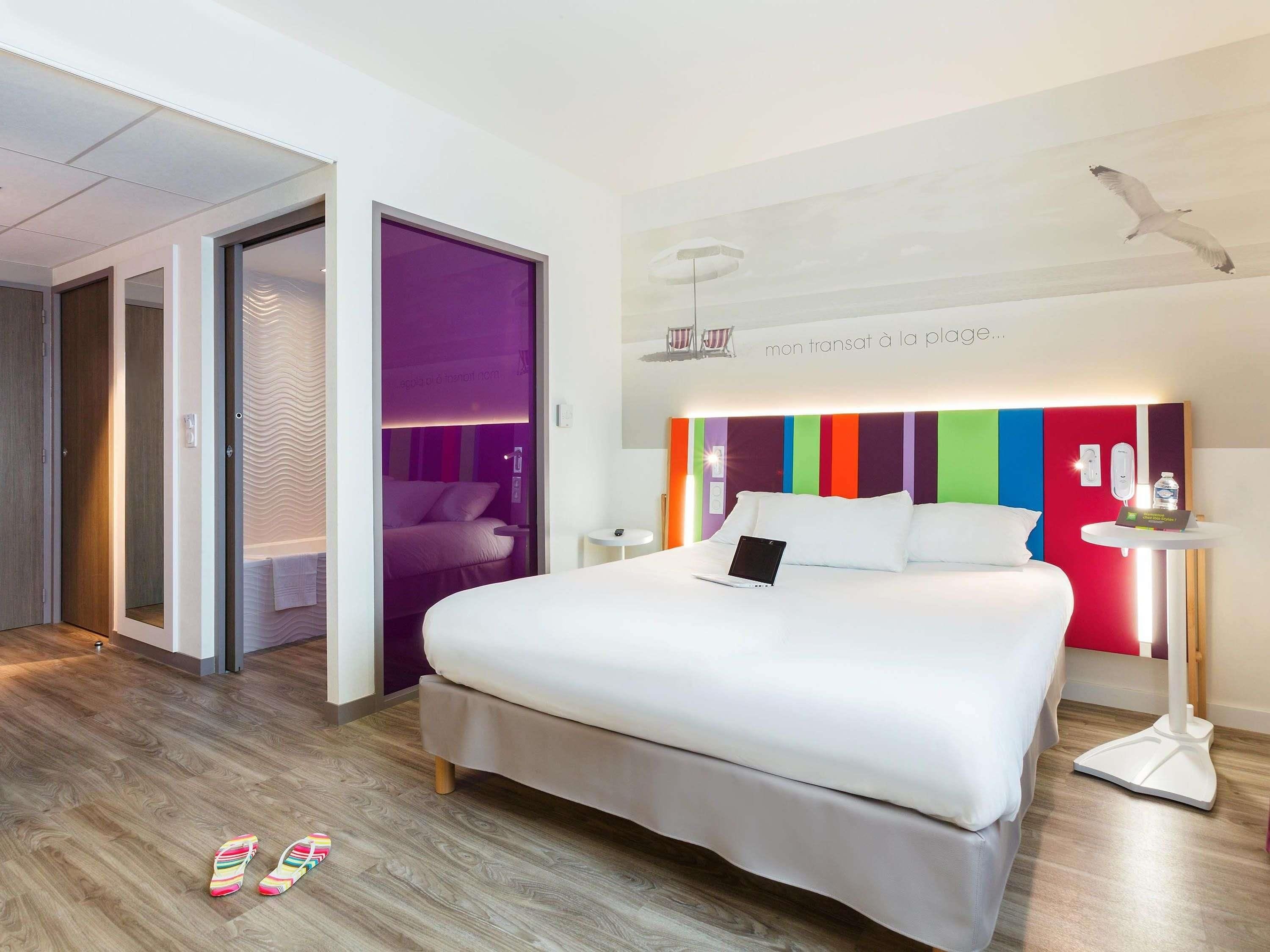 A modern hotel room with a large bed, a desk with a laptop, a bedside table with a phone and water bottle, and a colorful headboard.