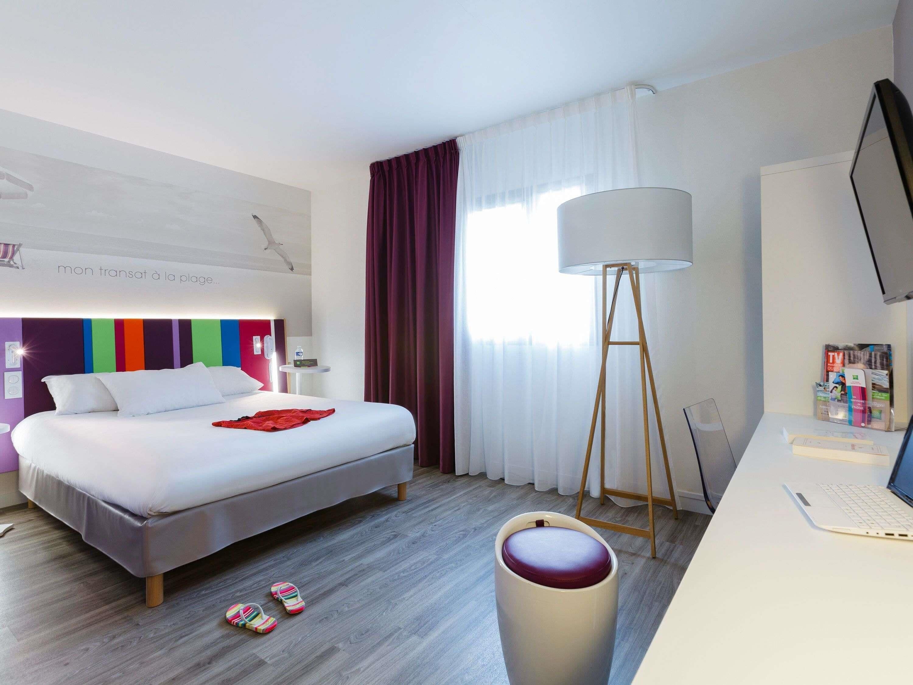 A hotel room with a bed, a desk with a laptop, a floor lamp, and a window with curtains.
